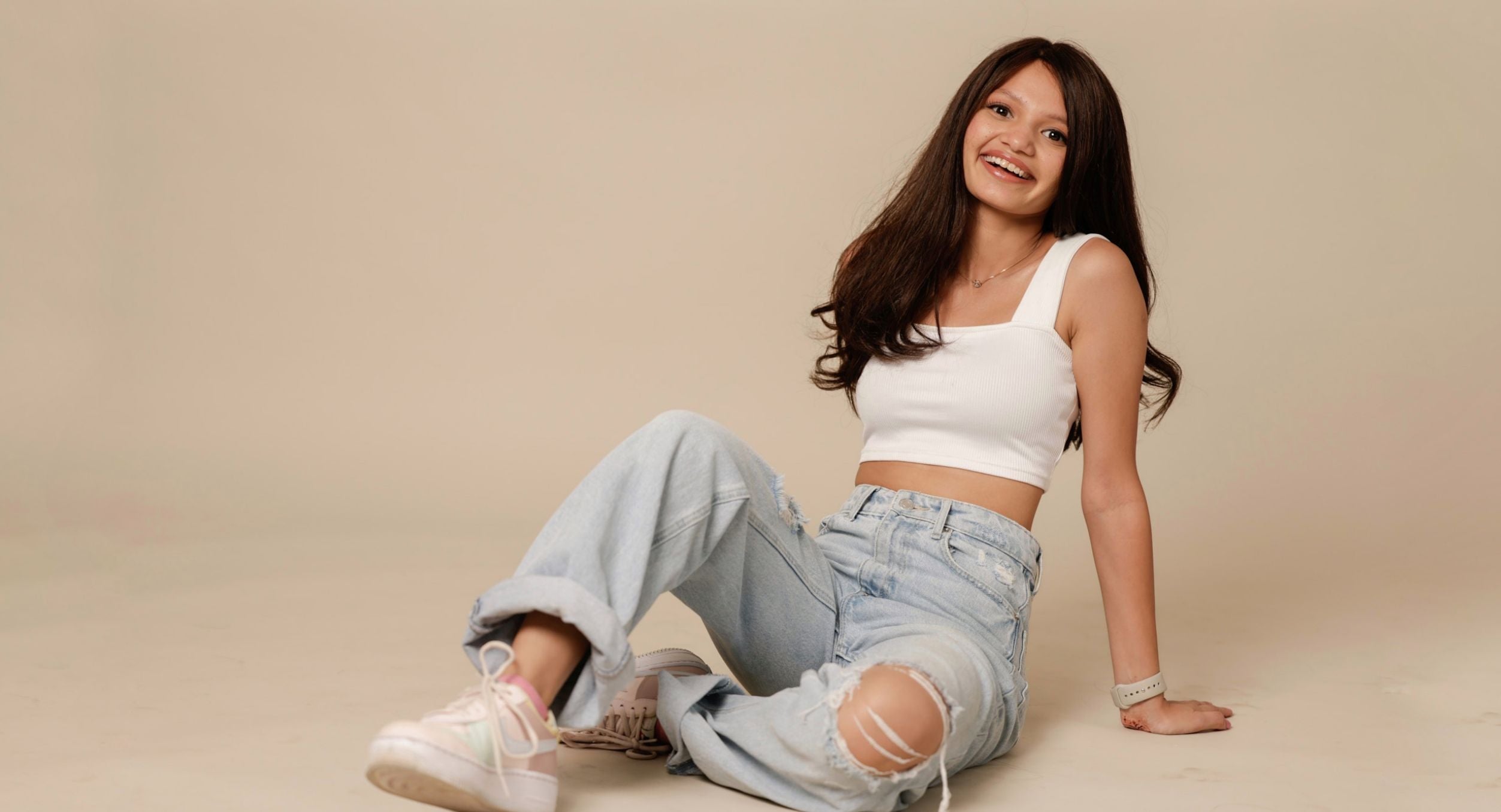 A smiling woman with long hair sits on the floor, wearing a crop top and ripped jeans.